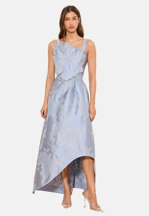 Moteris dėvi lengvą mėlynos spalvos gėlėtą suknelę be rankovių su asimetriniu kraštu ir dideliu audinio gėlės dekoru krūtinės srityje, derina ją su aukštakulniais sandalais.