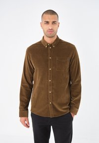 Cămașă din corduroy brun cu guler tip button-down, buzunar pe piept și nasturi de culoare aurie. Mâneci lungi cu manșete cu nasturi.