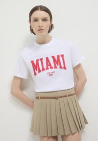 Frau in weißem T-Shirt mit rotem Aufdruck "MIAMI EST. 1987 U.S.A" und beiger plissierter Rock mit einem dünnen braunen Gürtel, stehend vor weißem Hintergrund.