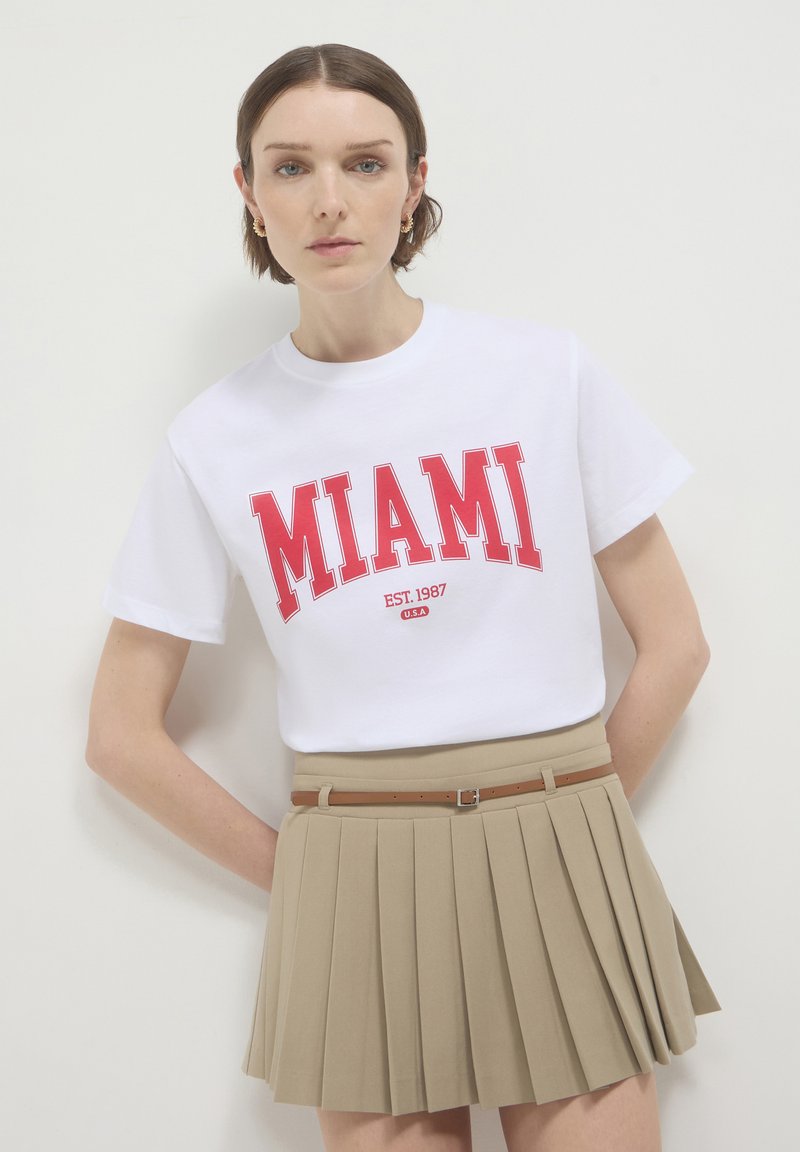 Frau in weißem T-Shirt mit rotem Aufdruck "MIAMI EST. 1987 U.S.A" und beiger plissierter Rock mit einem dünnen braunen Gürtel, stehend vor weißem Hintergrund.
