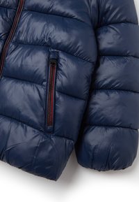 Giacca puffer blu navy con design trapuntato, texture liscia, dettagli della cerniera rossi e tasca laterale con zip.