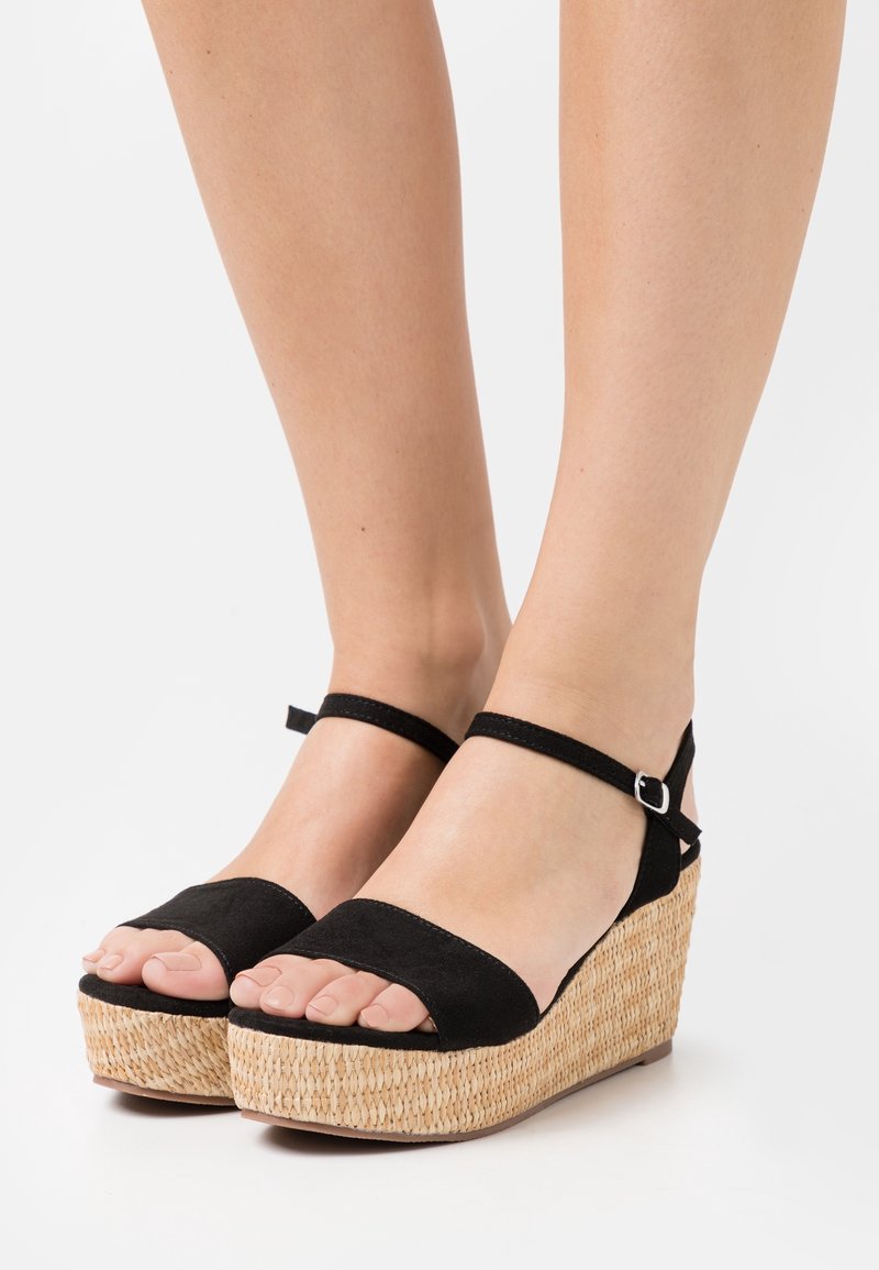 Sandales à talon compensé en paille tissée, brides en daim noir traversant les orteils et les chevilles, portées pieds nus sur des jambes à peau claire.
