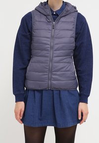 Gepolsterte ärmellose Weste in Dunkelgrau mit Kapuze, ausgestattet mit einem Frontreißverschluss und gestepptem Design, getragen über einem blauen Langarmshirt und einem Rock.