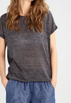 Woman wearing a loose dark gray short-sleeve top and blue casual pants with hands in pockets, standing against a plain background.