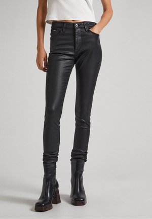 Femme portant un pantalon skinny en cuir noir et des bottines noires à talons, debout avec une main dans la poche sur fond gris clair.