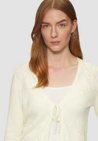 Cardigan en crème avec un col en V, fermeture à nouer sur le devant et motif en forme de cœur texturé. Matière tricotée légère avec des manches longues.