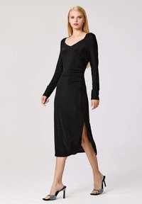 Femme portant une robe midi noire à manches longues avec une fente sur le côté, assortie à des sandales à talons ouverts noires ornées de détails.