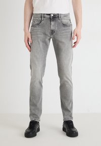 Szürke farmernadrág, slim fit szabással, öt zsebbel és finom kopással. Fekete sportcipőkkel párosítva, világos háttérrel.