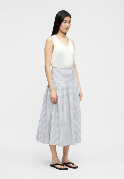 Femme aux longs cheveux noirs portant un haut blanc sans manches, une jupe midi rayée gris clair taille haute, et des tongs noires, debout de profil.