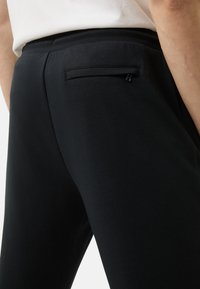 Czarne spodnie o gładkiej fakturze i elastycznym pasku w talii; posiadają tylnią kieszeń z czarnym zamkiem błyskawicznym oraz subtelne logo.