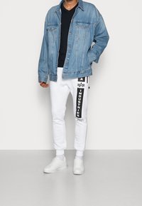 Giacca di jeans azzurro chiaro, maglietta nera con scollo a V, pantaloni della tuta bianchi con stampa del logo nero e sneakers bianche. Look streetwear casual.