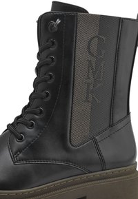 Bottine noire à lacets, présentant des accents en tissu texturé et un logo embossé sur un panneau gris, affichant une semelle en caoutchouc robuste.