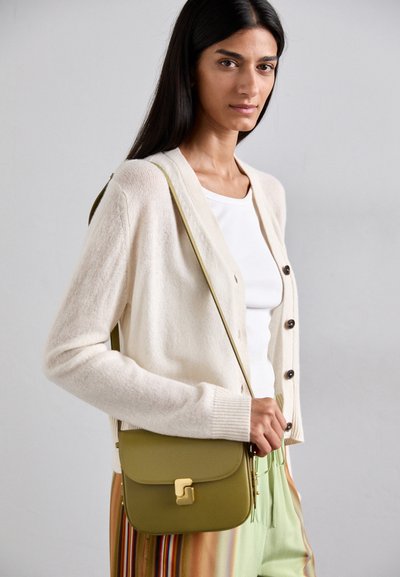 Olīvzaļš crossbody soma ar noapaļotu formu, zelta toņa detaļām un kustamo vāku, apvienota ar krēmkrāsas cardigan un baltu topu.