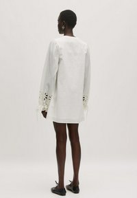 Weißes Leinenkleid mit langen Puffärmeln, die mit Spitzenbesatz und Bändern an den Manschetten verziert sind. Gerader Schnitt mit einem schlichten Rückendesign.