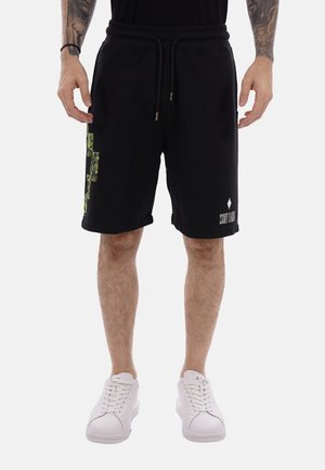 Man wearing black knee-length shorts with green graphic on side and white sneakers, standing with arms relaxed.