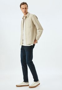 Camisa chaqueta beige abotonada con dos bolsillos en el pecho, llevada sobre una camisa blanca, combinada con jeans oscuros y zapatillas blancas con detalles en marrón.
