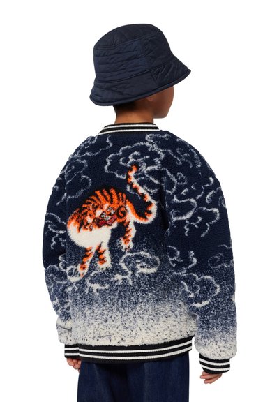 Veste en peluche bleu marine et blanche avec une surface texturée, présentant un design de tigre coloré au dos et des poignets et un ourlet rayés.