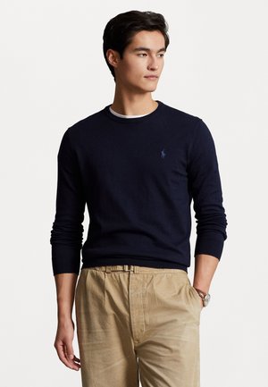 Navy crew-neck sweater made of knit fabric, featuring a small embroidered logo on the chest, paired with light beige trousers.