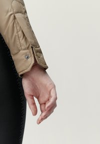 Padded, beige jackärm med tryckknappsstängning. Mjuk textur, med ett svart prickigt mönster nedanför, där en hand försiktigt vilar.