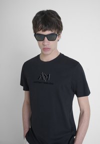 Black cotton t-shirt featuring a logo with silver accents. Model wearing angular black sunglasses against a neutral background.