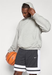Grå hættetrøje med løs pasform og ribbestrikket kant, parret med mørke shorts med hvide og sorte striber, mens der holdes en orange basketball.