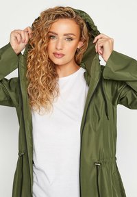 Giacca con cappuccio verde oliva con zip frontale, vita regolabile con cordino e materiale resistente all'acqua. Indossata sopra una T-shirt bianca.