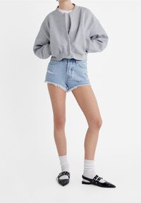 Persona que lleva una chaqueta bomber gris con botones, una camiseta blanca, shorts de denim desgastados, calcetines blancos y zapatos planos negros puntiagudos con correas.