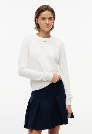 Giovane donna che indossa un maglione bianco a maniche lunghe con un piccolo logo e una gonna plissettata blu navy, in piedi contro uno sfondo semplice.