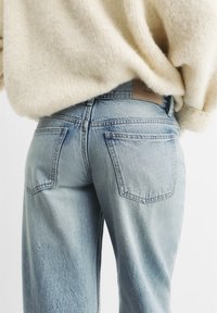 Jeans di denim azzurro chiaro con un lavaggio sbiadito, due tasche posteriori e un'etichetta in pelle marrone sulla cintura. Indossati con un maglione color crema.