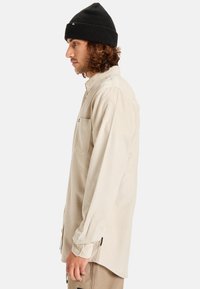 Camisa de pana beige con cuello tipo button-down, mangas largas y un bolsillo en el pecho; usada con un gorro de punto negro.