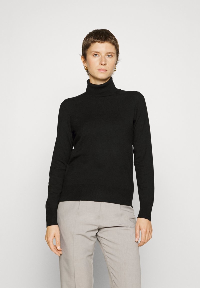 Pull à col roulé noir en tissu lisse, doté de manches longues et d'une silhouette ajustée, associé à un pantalon gris clair.