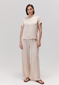 Beżowa jedwabna bluzka z krótkim rękawem o luźnym kroju, zestawiona z szerokimi spodniami. Miękka tekstura, minimalistyczny design i płaskie sandały dopełniają całość.
