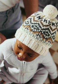 Bonnet pour enfant tricoté en crème avec des accents à motifs en vert et marron, surmonté d'un pompon en fourrure. Bande côtelée à la base.