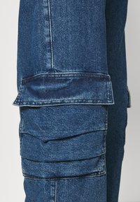 Blue denim cargo pants featuring layered pockets, a textured fabric, and a loose-fit design with visible stitching details.