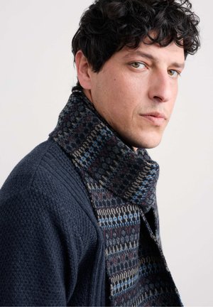 Jūros laivyno vilnos kardiganas su tekstūruotu mezgimu ir raštuotu šaliku, kuriame dominuoja tamsiai mėlyni, rudi ir pilki raštai. Artimas, vieno profilio kadrė.