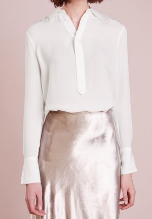 Kobieta w białej bluzce z długim rękawem, włożonej w błyszczącą metaliczną, jasnoróżową spódnicę, stojąca na tle bladoróżowym.