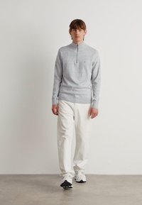 Pulover gri deschis cu fermoar, cu guler înalt, combinat cu pantaloni albi lejeri și adidași albi cu accente negre.