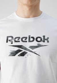 Bílý bavlněný tričko s velkým, opotřebovaným černým logem Reebok a ostrými grafickými prvky na hrudi. Krátké rukávy, kulatý výstřih.