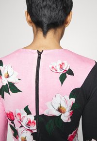 Personne avec des cheveux courts et foncés portant un haut à motifs floraux rose et noir avec une fermeture éclair noire visible dans le dos.