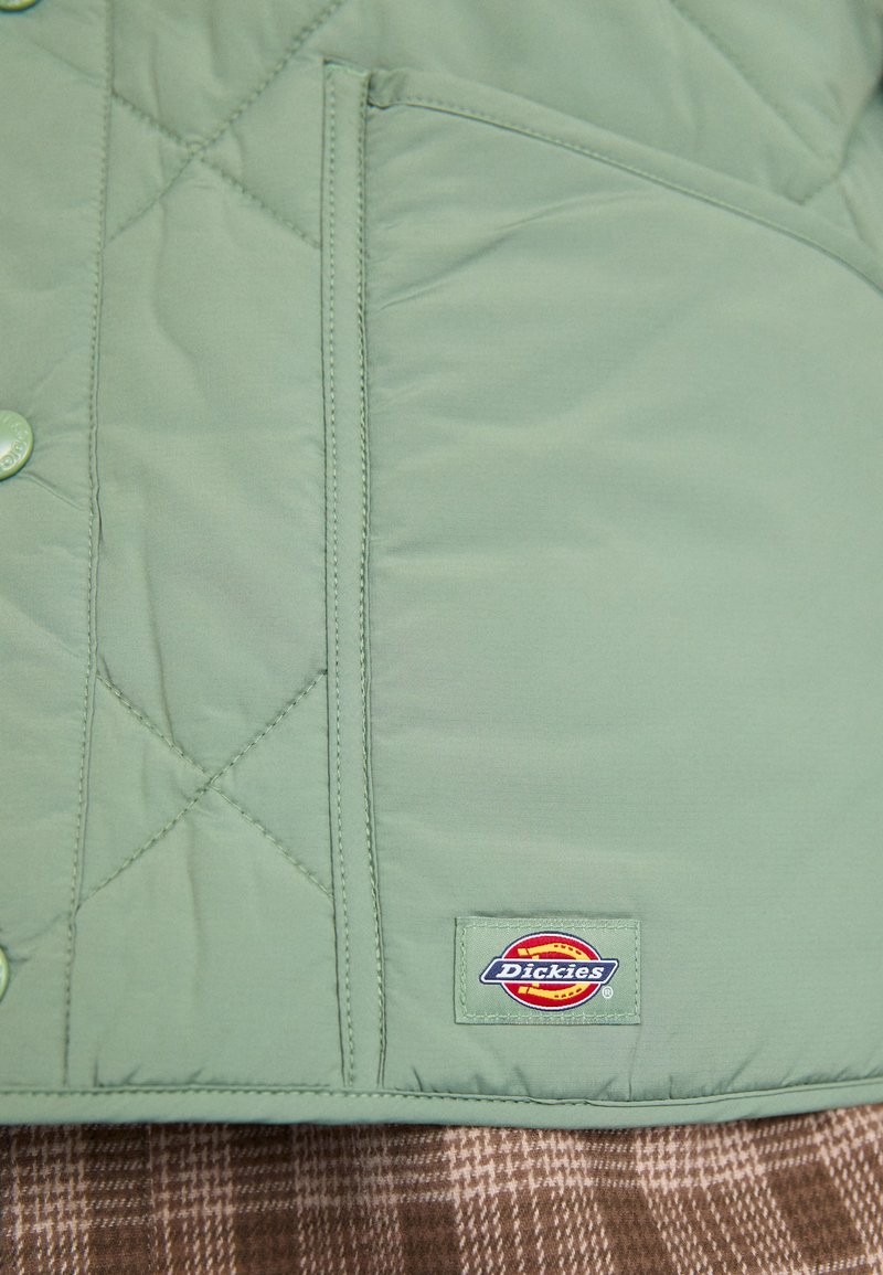 Quiltet grøn jakke med kuppelformede knapper, en lomme og et Dickies-logo. Fremstillet af glat, let stof.