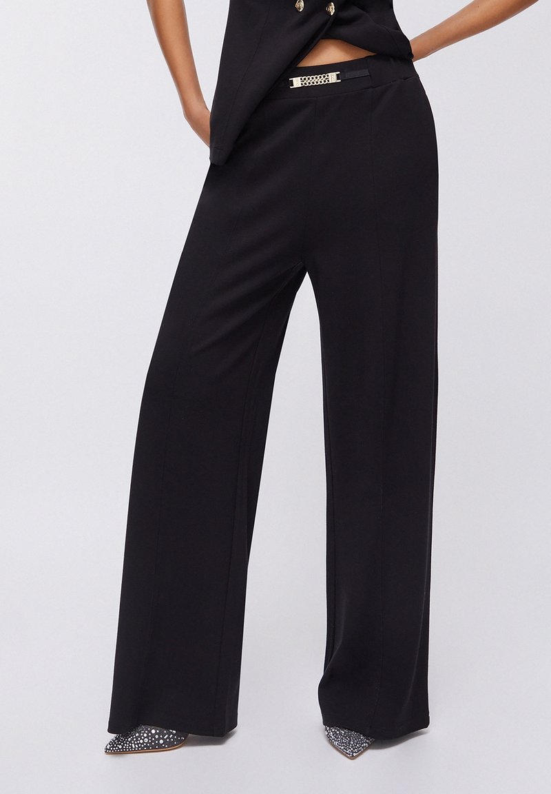 Femme portant un pantalon noir à jambes larges taille haute avec un détail de ceinture dorée et des chaussures pointues ornées de strass sur un fond uni.