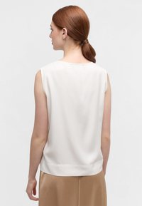 Mouwloze, crèmekleurige blouse met een gladde textuur en een ronde halslijn. De achterkant is eenvoudig met een subtiele zoom en is gecombineerd met een beige rok.