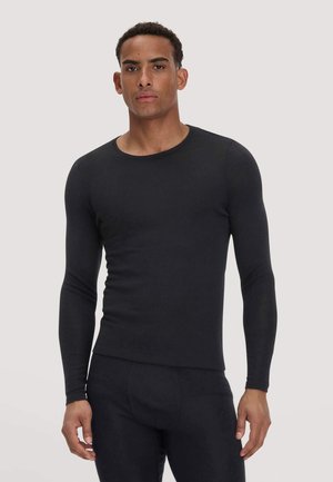 Mann steht frontal, trägt ein eng anliegendes schwarzes Langarmshirt und passende schwarze Hose vor einem einfarbigen hellen Hintergrund.