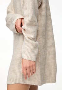 Robe-pull beige en maille avec une texture côtelée, des manches amples et une coupe décontractée, montrant une main saisissant le poignet de la manche.