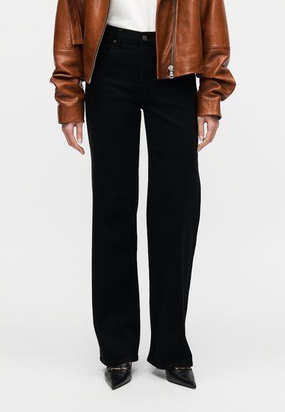 Μαύρο παντελόνι με φαρδιά πόδια από λείο ύφασμα, συνδυασμένο με εφαρμοστή λευκή πουκάμισα και καφέ δερμάτινο μπουφάν. Τα μυτερά μαύρα παπούτσια ολοκληρώνουν την εμφάνιση.