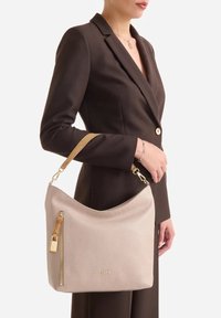 Donna in blazer marrone scuro che tiene una borsa in pelle beige con cerniera dorata e tracolla marrone chiaro appoggiata al braccio.
