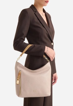 Donna in blazer marrone scuro che tiene una borsa in pelle beige con cerniera dorata e tracolla marrone chiaro appoggiata al braccio.