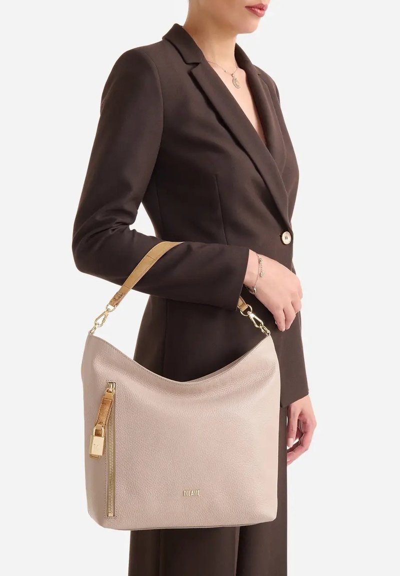 Donna in blazer marrone scuro che tiene una borsa in pelle beige con cerniera dorata e tracolla marrone chiaro appoggiata al braccio.