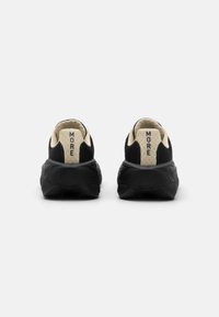 Chaussures de sport noires avec un dessus en tricot texturé, des accents de talon crème et "MORE" imprimé à l'arrière. Semelle douce avec une semelle à motif.