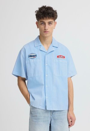 Hellblaues kurzärmeliges Hemd im entspannten Stil, mit zwei Fronttaschen und bestickten Logodetails auf der Brust. Weiche Textur.